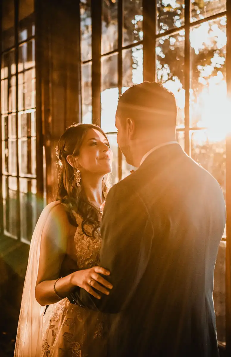 Foto de matrimonio parado junto a ventana por la que entran los rayos de sol